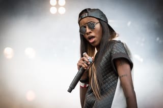 Angel Haze in a backwards leather cap and round mirrored sunglasses, rapping into a handheld microphone on a festival stage