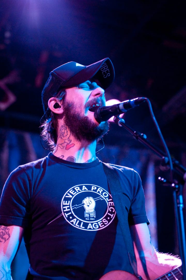 Ben Bridwell, bearded, in a cap and a dark T-shirt, singing into a microphone on stage