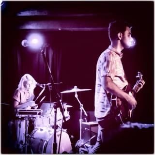 Big Scary on a small stage under purple light, a drummer behind the kit on the left and a guitarist in a patterned shirt on the right