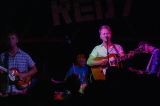 Four musicians on a dark stage under a large RED 7 sign: a bassist and a singer with an acoustic guitar at microphones, a drummer in a cap between them and a guitarist on the right