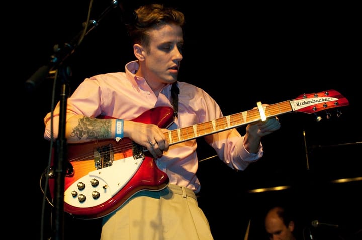 Christopher Owens in a pink shirt playing a red Rickenbacker guitar on a dark stage