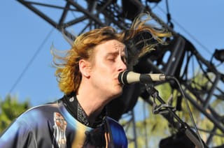 Christopher Owens singing into a microphone outdoors, his long blond hair blowing in the wind