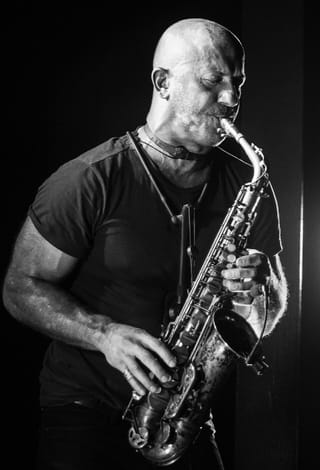 Black-and-white photo of Colin Stetson, eyes closed, playing a saxophone on a dark stage