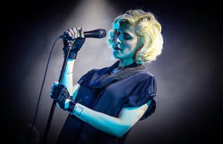 Dessa singing into a microphone on a stand under a blue spotlight