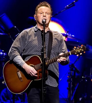 Don Henley singing into a microphone and playing an acoustic guitar on stage