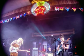 Two guitarists singing at microphones on a stage under a neon star sign and strings of bunting, with a drummer between them