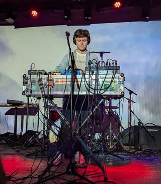 Warren Hildebrand standing behind a table of electronic equipment and cables on a stage lit red and blue
