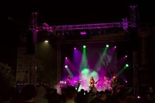 A band playing on an outdoor festival stage under purple and green lights, seen from the back of the crowd