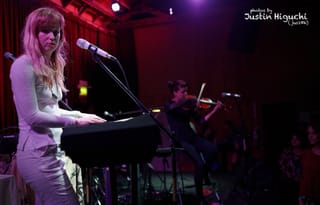 Gracie and Rachel on a small red-lit stage, a blonde singer at a keyboard in the foreground and a violinist seated behind her