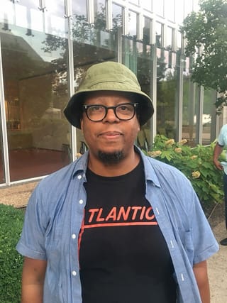 Jeff Parker in a green bucket hat, glasses and a denim shirt, standing in front of a glass building