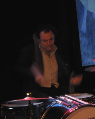 Jim White playing a drum kit on a dark stage