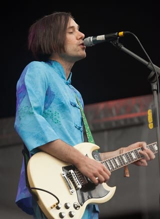 Kevin Barnes in a pale blue patterned tunic, eyes closed, singing into a microphone while playing a cream electric guitar