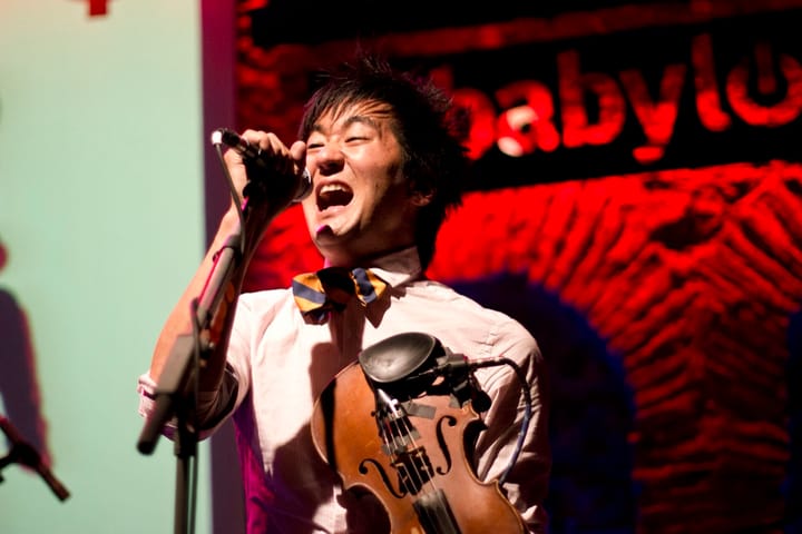 Kishi Bashi singing into a handheld microphone with a violin under his arm, in a striped bow tie, in front of a red backdrop