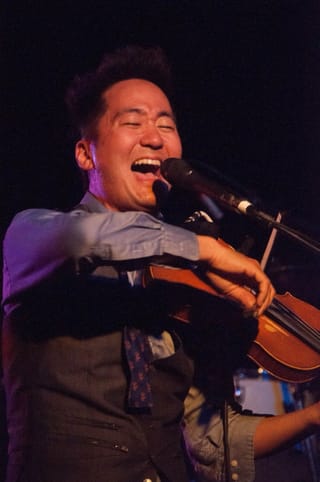 Kishi Bashi singing with his eyes closed into a microphone while playing violin, in a grey waistcoat and patterned tie