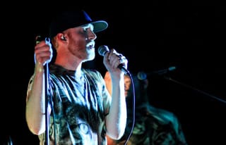 Logan Lynn singing into a handheld microphone on a dark stage, in a baseball cap and a printed T-shirt