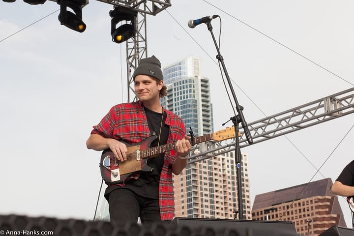 Mac DeMarco in a grey beanie and a red plaid shirt, smiling and playing an electric guitar on an outdoor stage, with tall buildings behind him