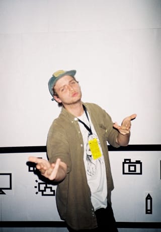 Mac DeMarco in a cap and an olive shirt, arms held out, posing against a white wall