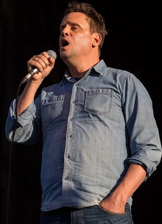 Mark Kozelek in a denim shirt singing into a handheld microphone on an outdoor stage