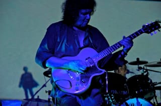Massimo Morante playing a white electric guitar on a stage lit blue