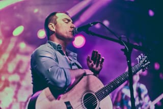 Matthew Tonner singing into a microphone and playing an acoustic guitar on a stage lit in pink and purple