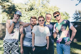 Five young musicians posing on grass in front of trees on a sunny day, a woman in a white mesh top in the centre