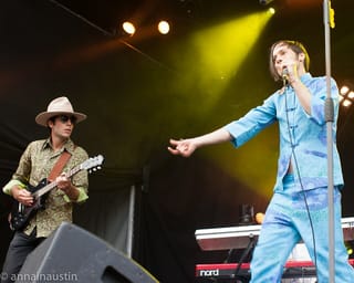 of Montreal on stage: the singer in a pale blue suit gesturing with a handheld microphone, a guitarist in a wide-brimmed hat and sunglasses on the left