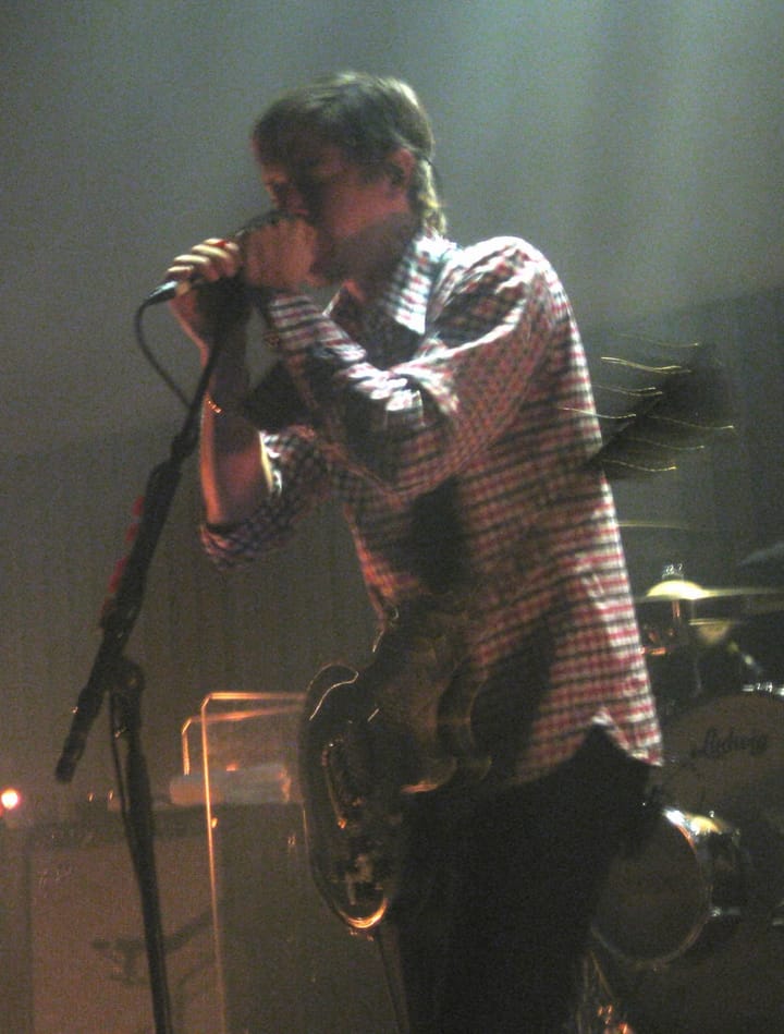 Paul Banks in a checked shirt singing into a microphone on a dim stage, a guitar at his waist