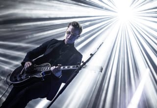 Paul Banks in a dark suit and glasses playing a black electric guitar against rays of white stage light