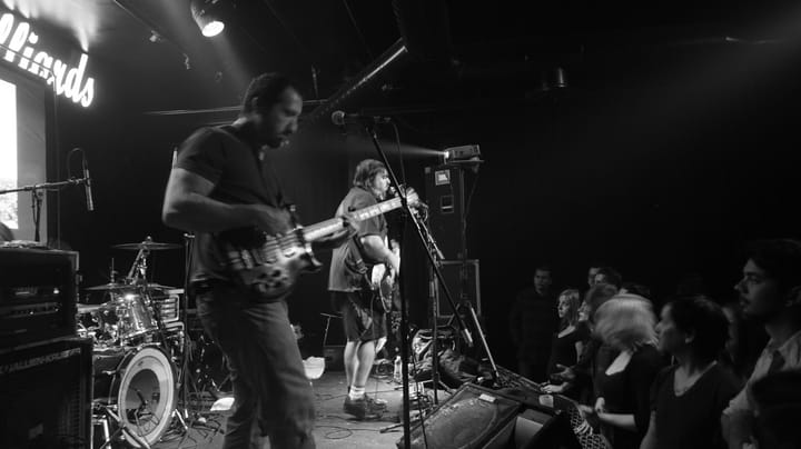 Black-and-white photo of a bassist in the foreground and a guitarist singing at a microphone behind him, on a club stage with a drum kit and a crowd at the edge
