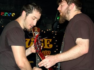 Two bearded musicians in brown T-shirts leaning together on a club stage, one bent over the headstock of a red guitar