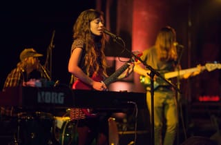 Quilt on a dark stage, a woman singing and playing a red electric guitar at the centre, a keyboard player on the left and a bass player on the right