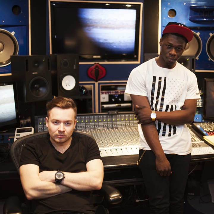 Two men at a large mixing console in a recording studio, one seated with arms crossed, one standing