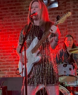 RAIGN singing into a microphone and playing a white electric guitar, in a fringed sequin dress, against a red-lit brick wall with a drummer behind her