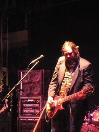 Rob Crow, long-haired and bearded in a dark jacket, looking down at a red electric guitar on a dim stage