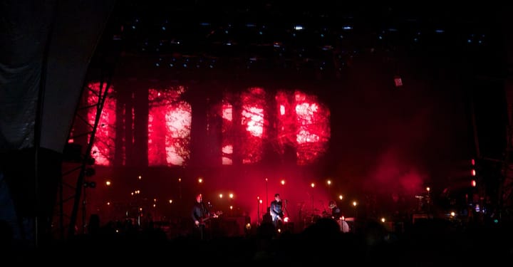 A band on a dark festival stage lit in red, with rows of small lamps on stands and a red projection behind them