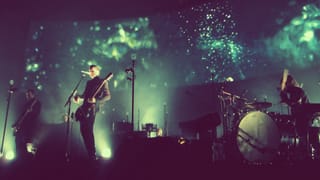 Three musicians on a dark stage in front of a green projected backdrop: a bassist, a singer with a guitar at the microphone, and a drummer