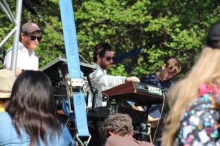 Three members of S U R V I V E at their keyboards on an outdoor stage, with trees behind them and the audience in the foreground
