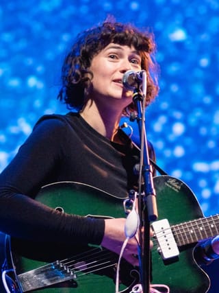 Kate Stables singing at a microphone with a green electric guitar, in front of a blue backdrop