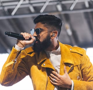 TOTEM singing into a handheld microphone on an outdoor stage, in sunglasses and a mustard suede jacket, his other hand on his chest