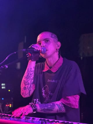 Trevor Powers singing into a handheld microphone with one hand on a keyboard, under purple stage light