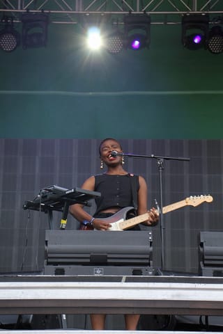 Vagabon singing and playing an electric guitar on a large outdoor stage, under a rig of stage lights