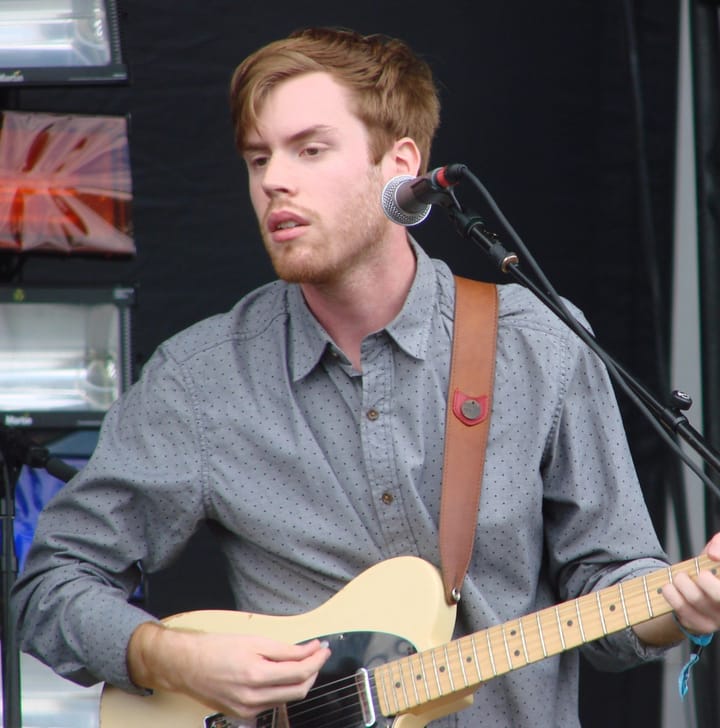 Jack Tatum in a grey shirt singing into a microphone and playing a cream-coloured electric guitar