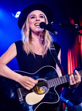 ZZ Ward smiling at a microphone in a wide-brimmed black hat, playing a black acoustic guitar under blue and red stage light